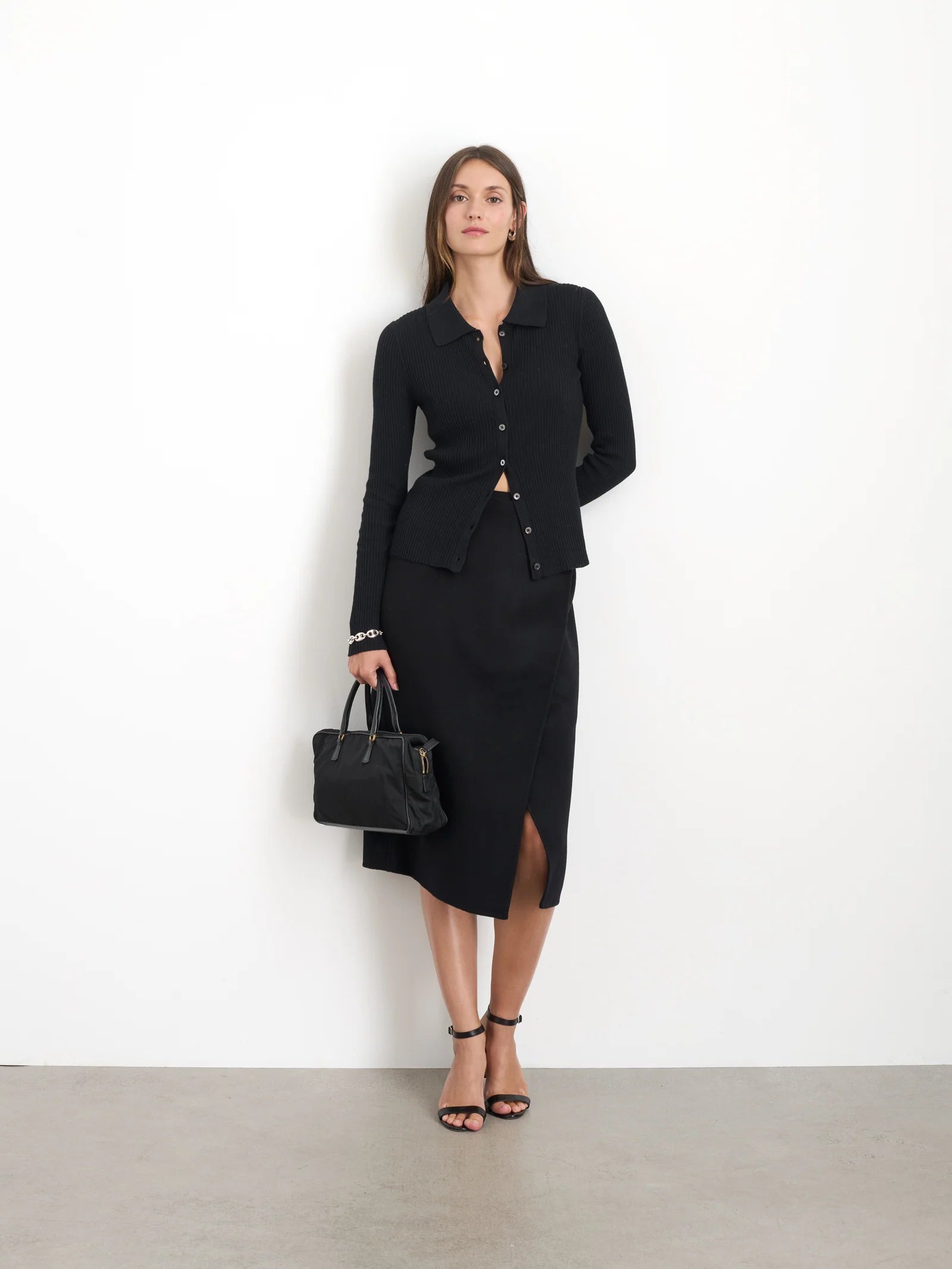 A woman wearing the Parker Midi Skirt by Alex Mill and a black button-up cardigan stands against a white background, perfectly dressed for holiday happy hours. She accessorizes with strappy heels and a black handbag, her long hair cascading loosely over her shoulders.