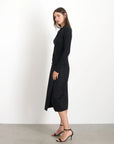 A woman with long brown hair stands sideways, dressed in a chic black long-sleeve top and the Parker Midi Skirt by Alex Mill, paired with high heels, ready for festive dinners or holiday happy hours. She is in a minimalist white room with a gray floor, exuding elegance and sophistication.