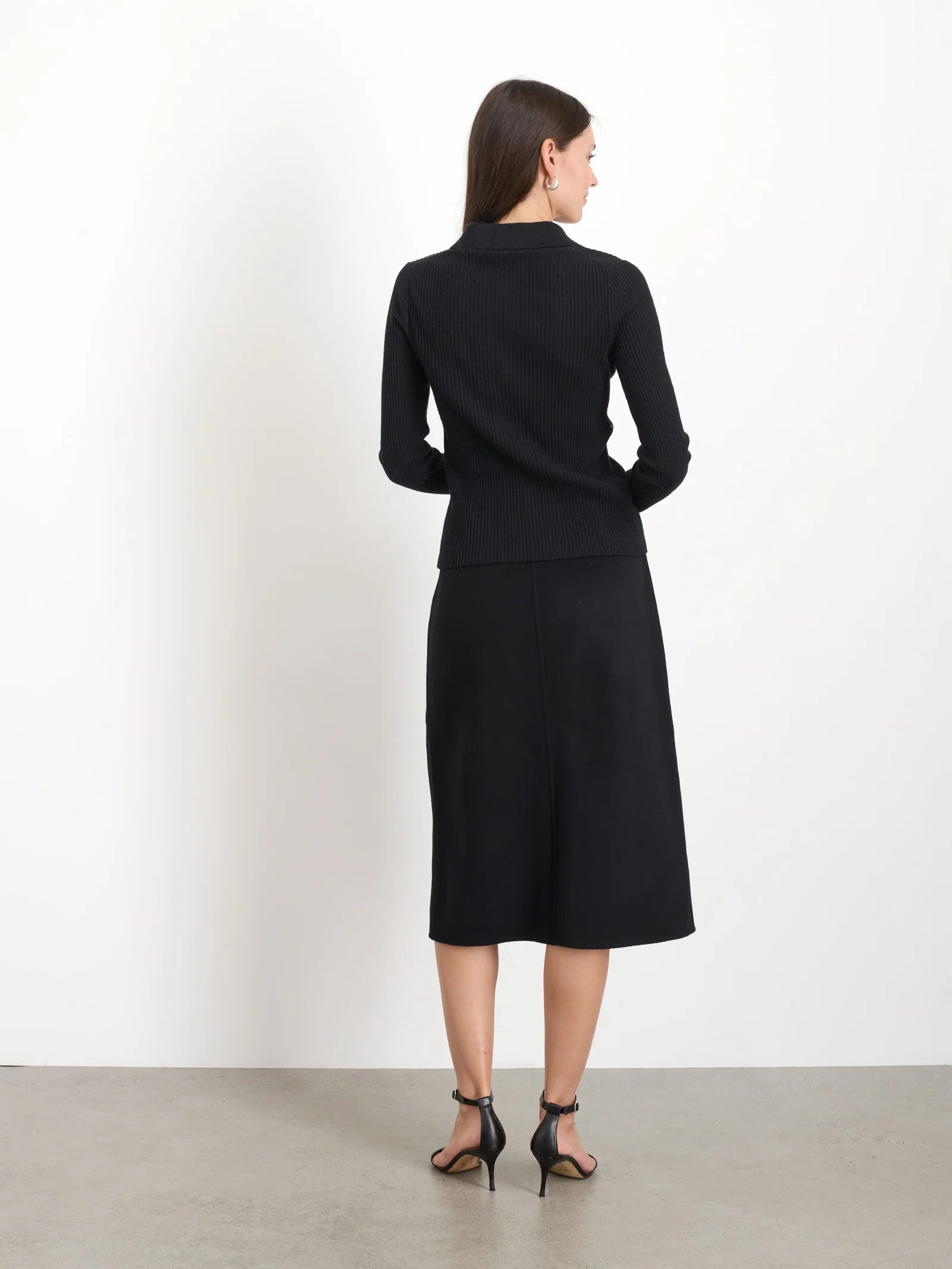 A woman with long dark hair stands with her back to the camera, poised for festive holiday gatherings. She is wearing a black long-sleeve top, the Parker Midi Skirt by Alex Mill, and black high-heeled sandals. The backdrop features a plain white wall and a gray floor, crafting an understated yet elegant scene.