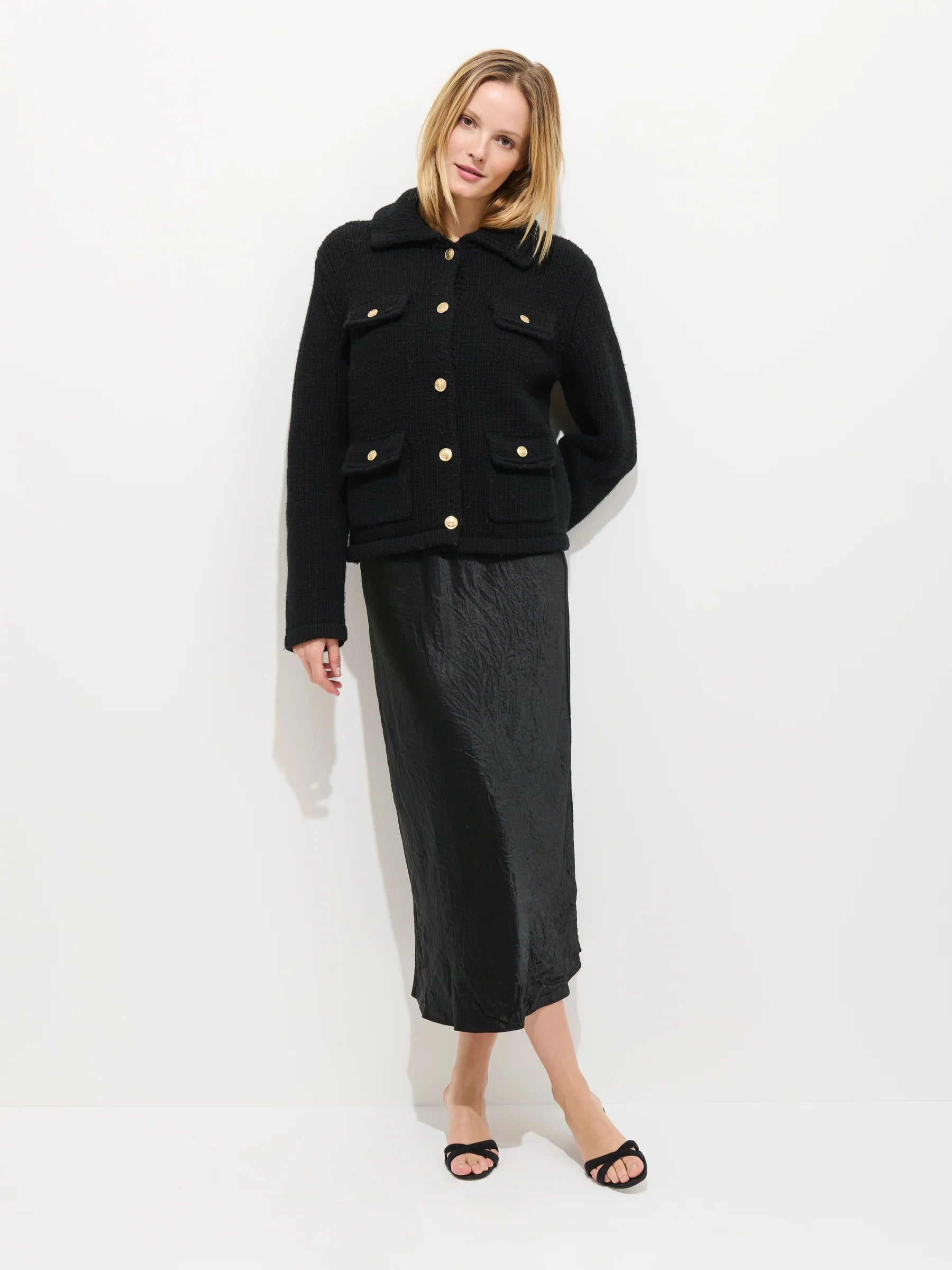 A woman with blonde hair poses casually against a white background wearing the Alex Mill Edinburgh Sweater, styled with a black midi skirt and open-toe sandals.