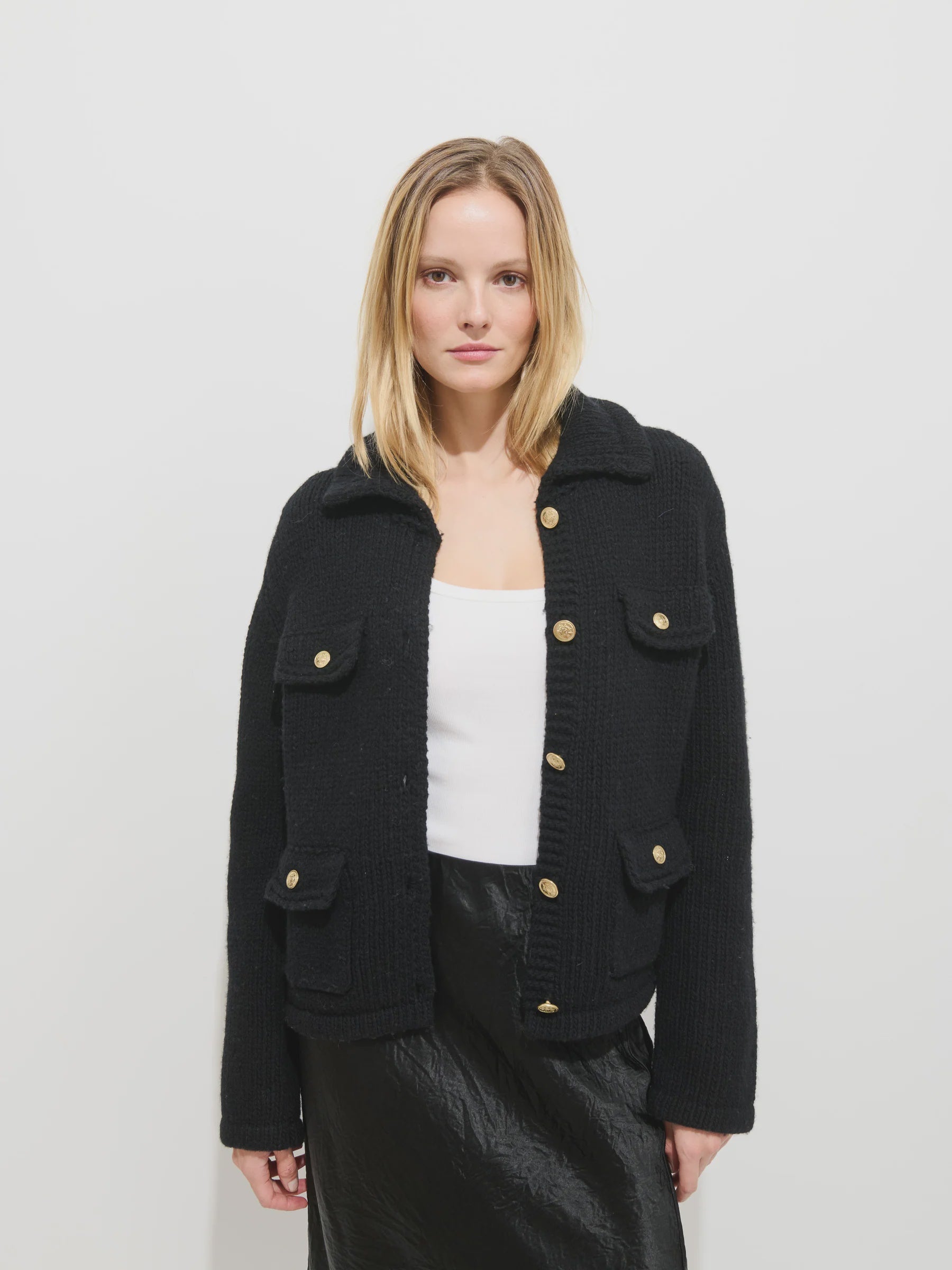 A woman with straight blonde hair wears the Edinburgh Sweater by Alex Mill, styled over a white top and black skirt, standing against a plain white background.