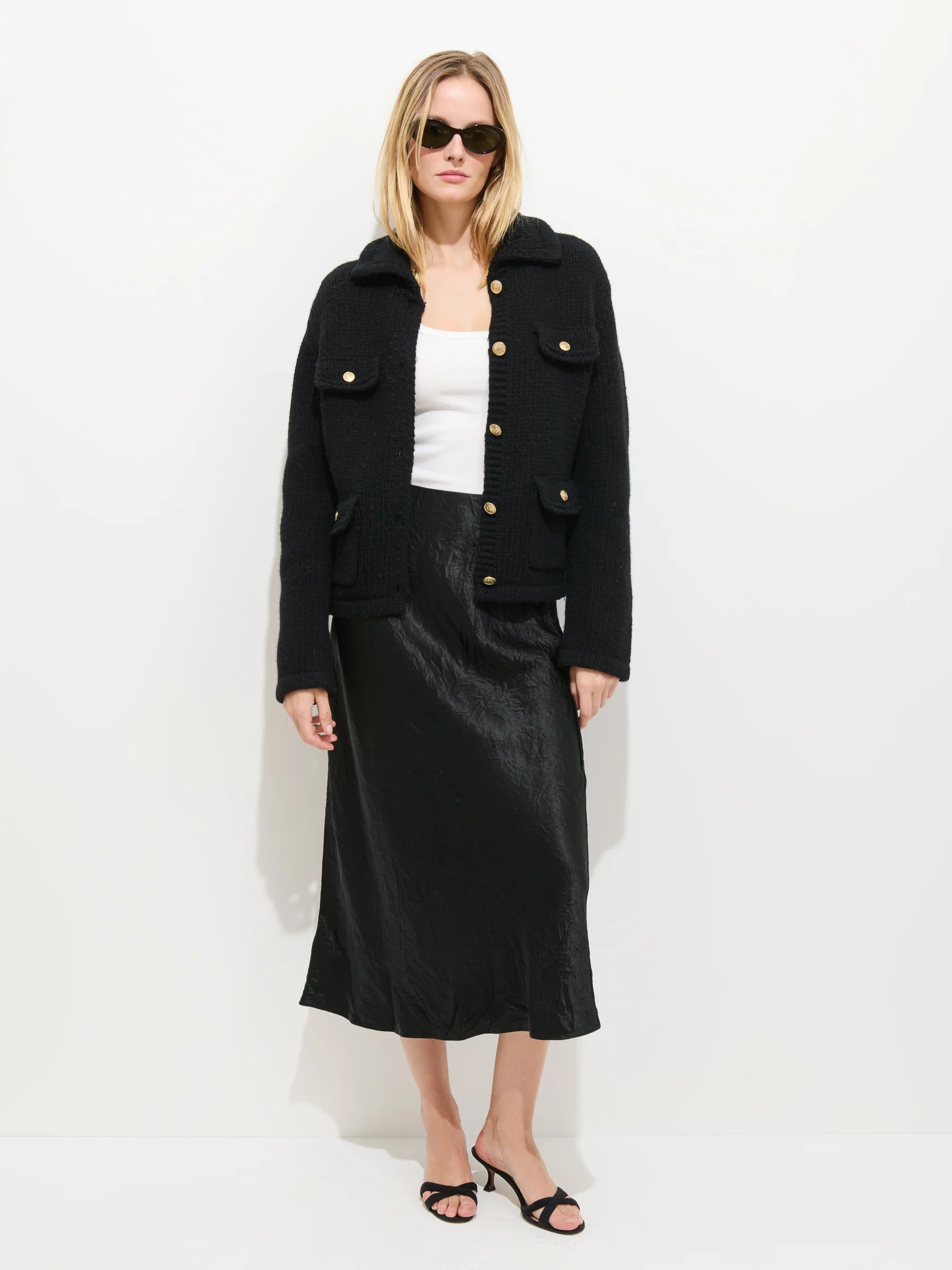 A woman wearing sunglasses, the Edinburgh Sweater by Alex Mill, a black satin midi skirt, and black heeled sandals stands against a plain white background.