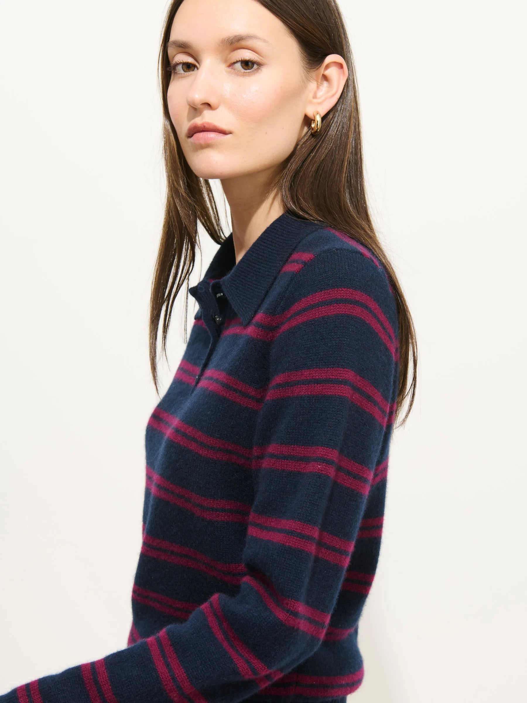 A woman with long brown hair wears the Alex Mill Alice Polo Cashmere, a luxury navy blue sweater with magenta stripes, and faces slightly to the side, looking at the camera against a plain white background.