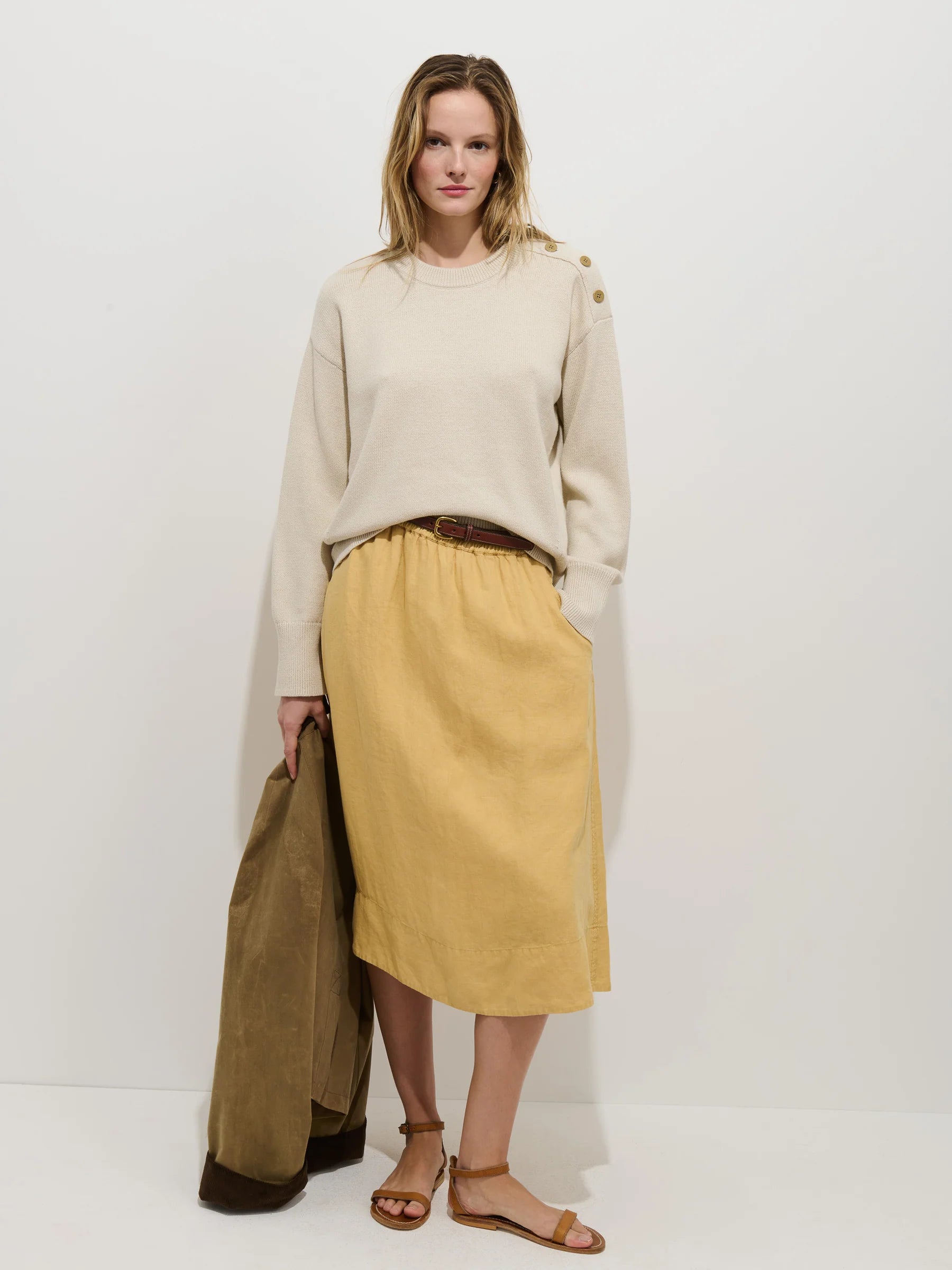 A woman stands against a white background wearing Alex Mill’s Maddie Skirt Linen in light yellow with pockets, a cream sweater with shoulder buttons, and brown sandals. She holds a brown jacket and looks at the camera.