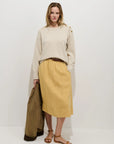 A woman stands against a white background wearing Alex Mill’s Maddie Skirt Linen in light yellow with pockets, a cream sweater with shoulder buttons, and brown sandals. She holds a brown jacket and looks at the camera.