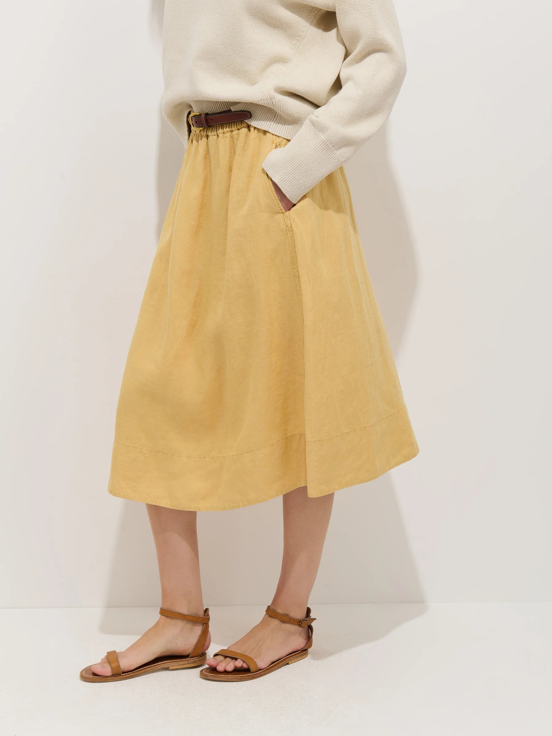 Model in a cream sweater tucked into the Alex Mill Maddie Skirt Linen—a loose, knee-length, garment-dyed skirt with pockets—styled with a brown belt and brown flat sandals, standing against a white background.