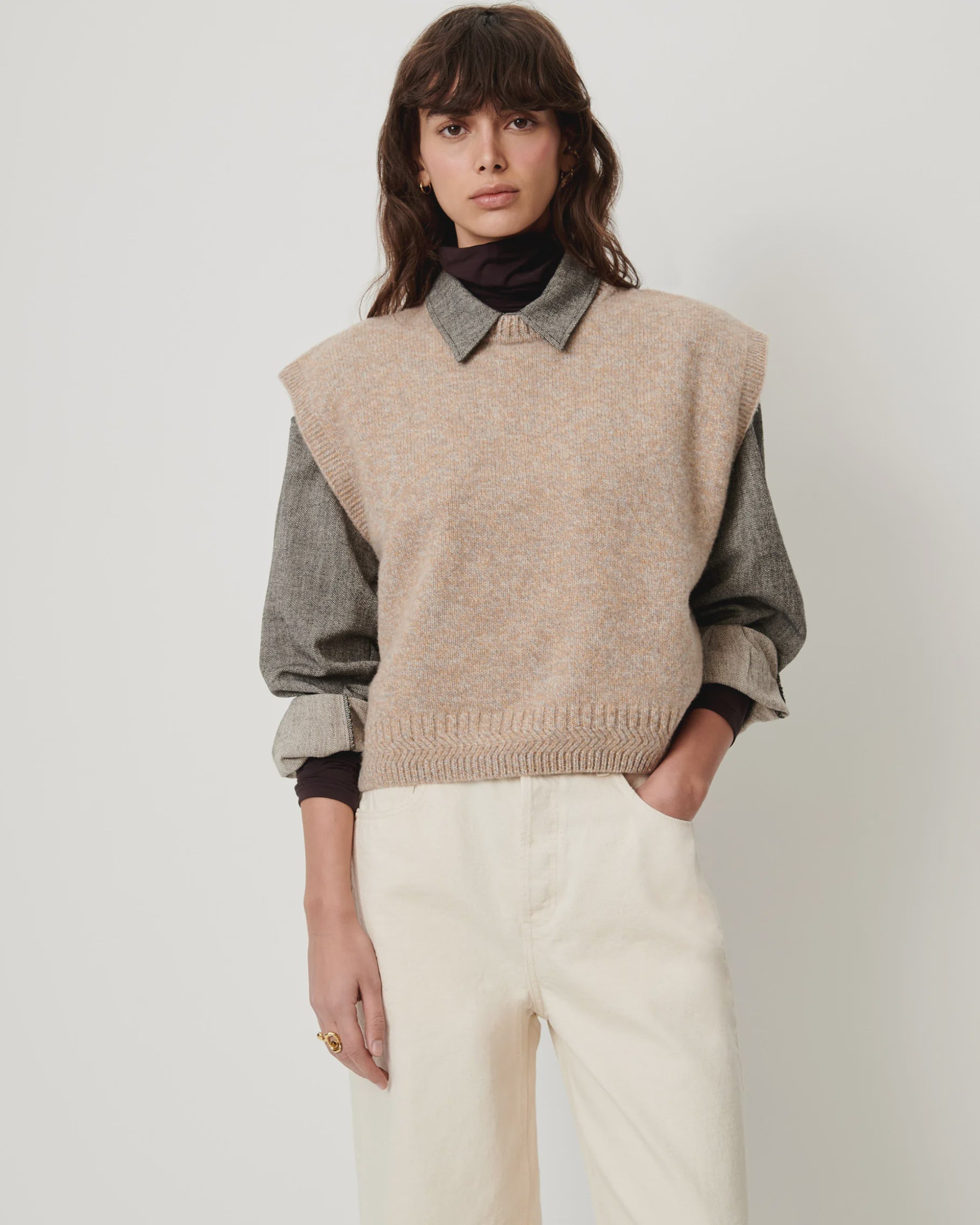 Sessun Fabinino beige sleeveless jumper layered over a gray shirt, styled with white pants for a relaxed look.