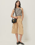 A woman stands against a white background wearing a sleeveless gray patterned top, Sessun Marco Bermuda Shorts in tan gabardine twill with pockets, black heeled shoes, and carries a small black crossbody bag.