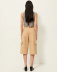 A person with long, dark hair stands facing away, wearing a sleeveless checked top, Sessun's Marco Bermuda Shorts in gabardine twill, and black loafers against a plain light background.