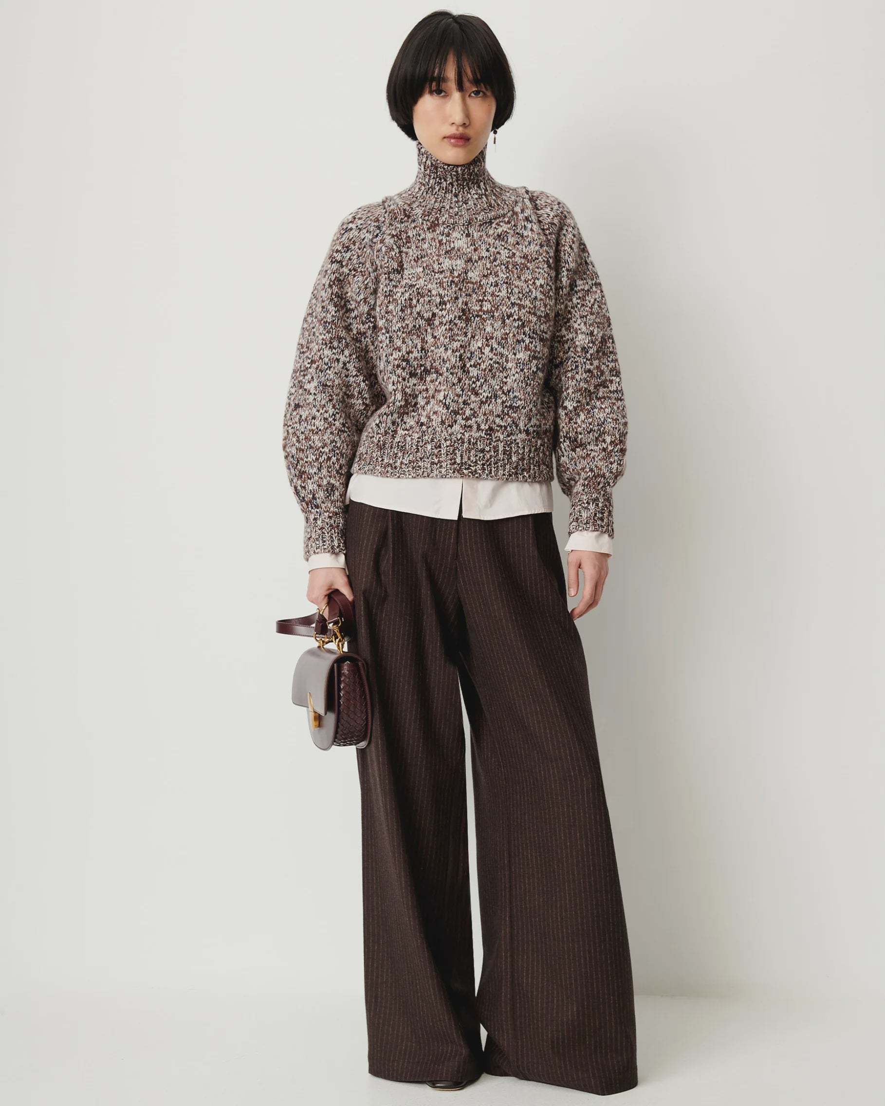 Sessun Orozco pleated large pants with a chunky knit turtleneck, styled by a white wall for a relaxed look.