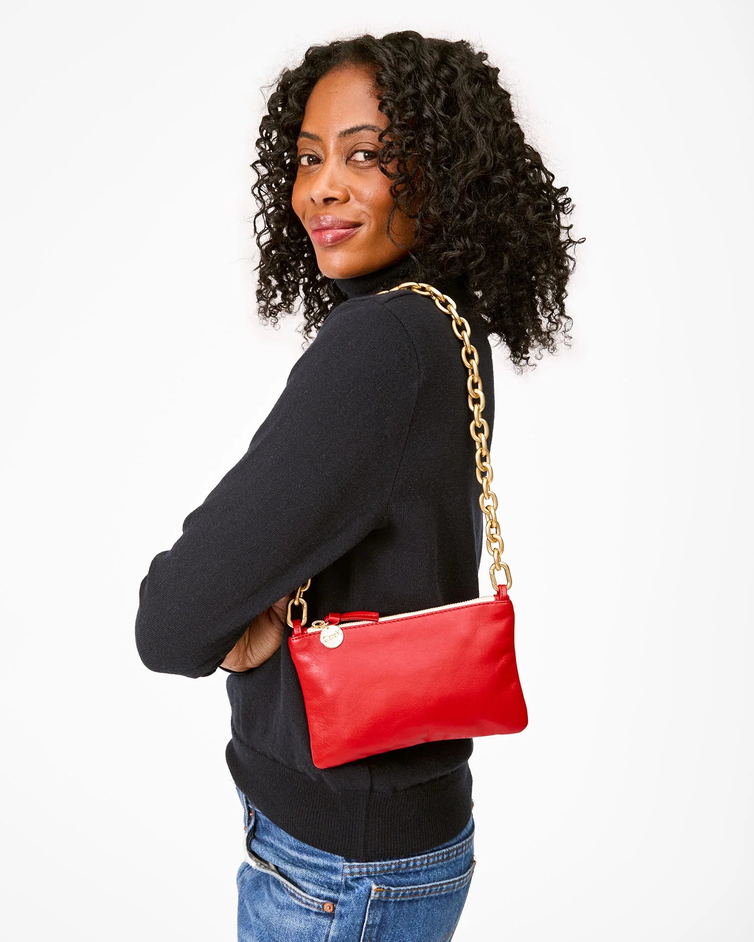 A woman with curly hair, wearing a black sweater and blue jeans, smiles while holding the Clare Vivier Wallet Clutch w/Tabs, crafted from Italian nappa leather with a gold chain strap. She stands against a white background, exuding elegance with her made-in-India style accessory.