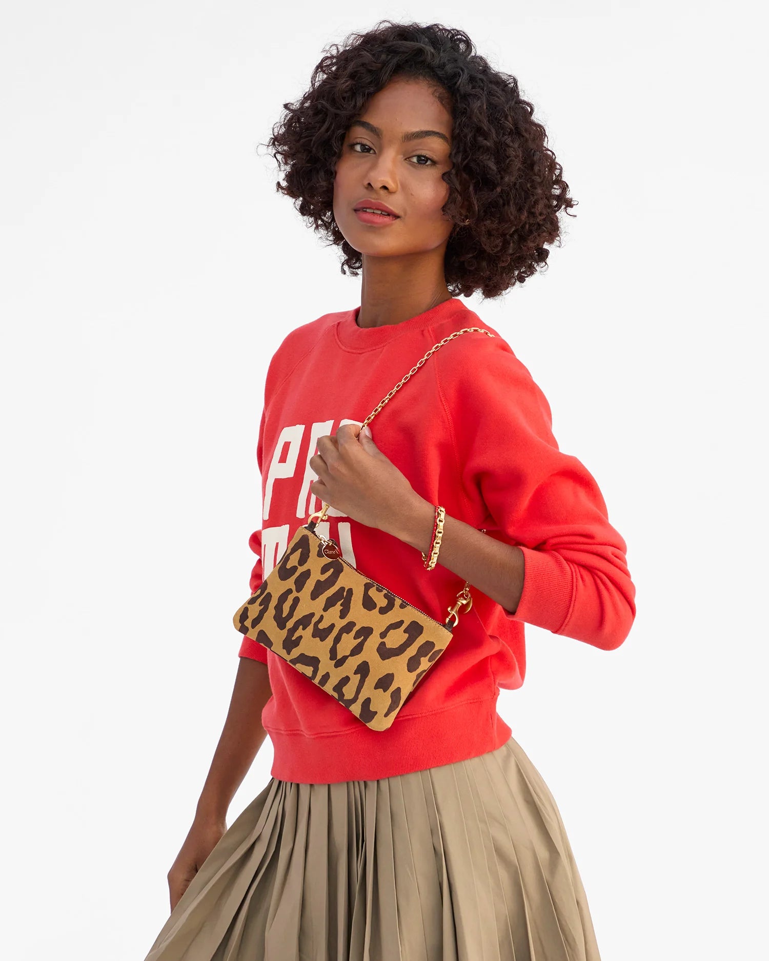 A woman with curly hair wears a red sweatshirt and beige pleated skirt, holding the Clare Vivier Wallet Clutch w/Tabs in Italian suede with a gold chain, standing against a plain white background.