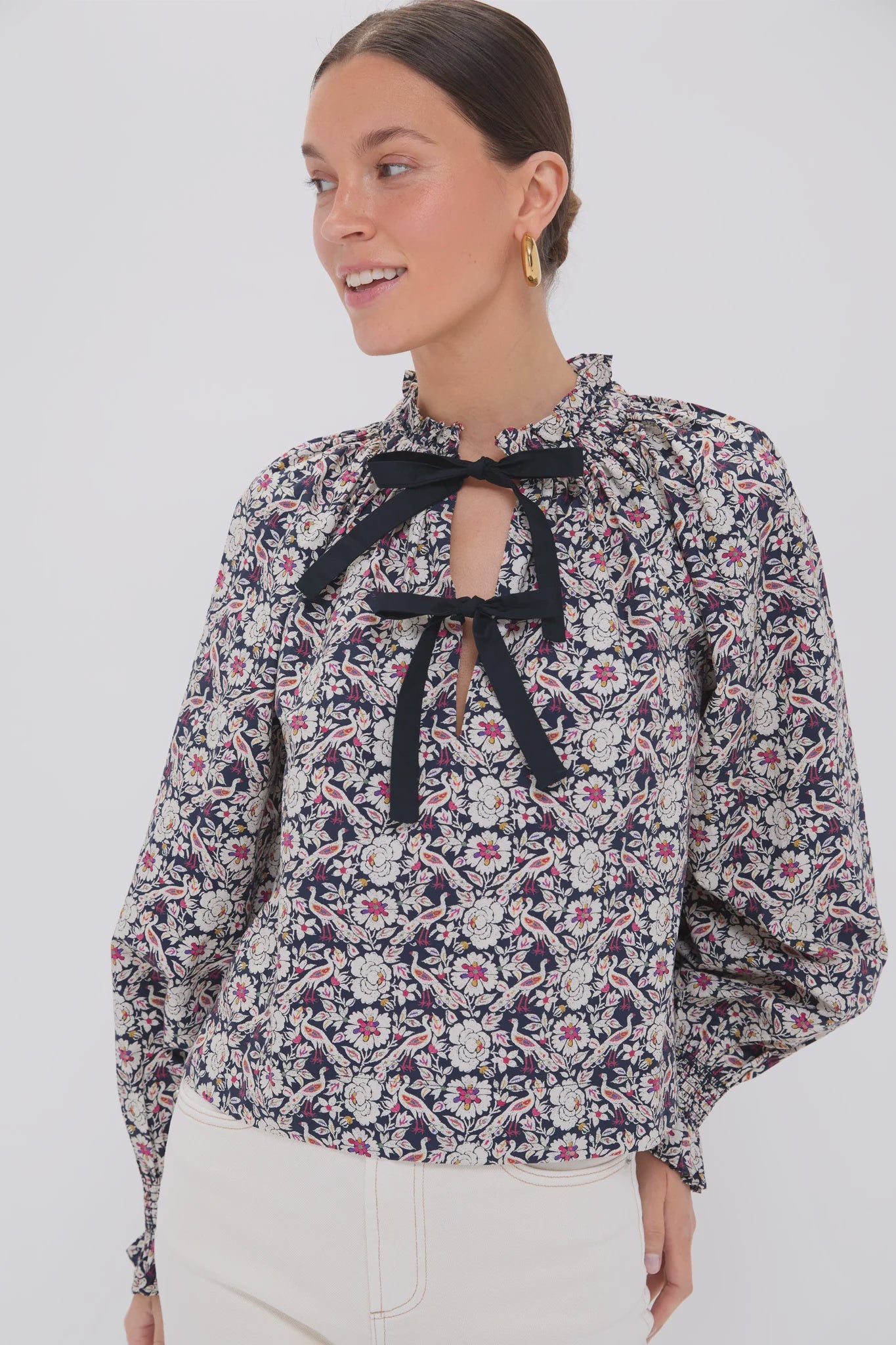 A woman wears the Rhode Aiden Top, featuring a pattern, keyhole, and a delicate ruffle collar. Paired with white pants and accessorized with gold hoop earrings and a black self-tie bow, she stands against a plain white background, looking aside with her hair pulled back, smiling.