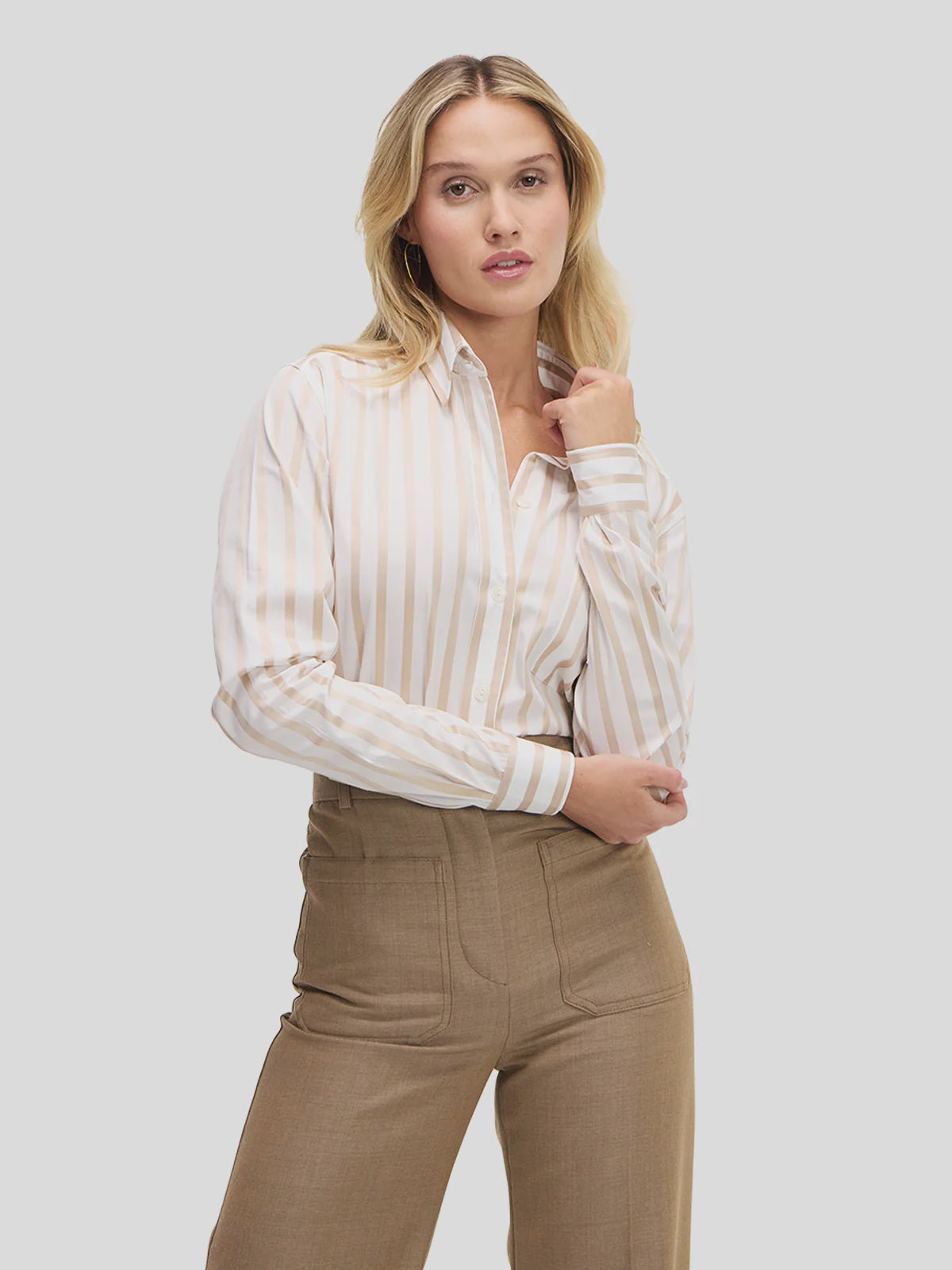 A woman with wavy blonde hair models the Weekend Shirt by Sarah Alexandra, LLC, featuring neutral stripes, tucked into high-waisted light brown pants. She poses against a plain background with one hand on her collar and the other on her hip.