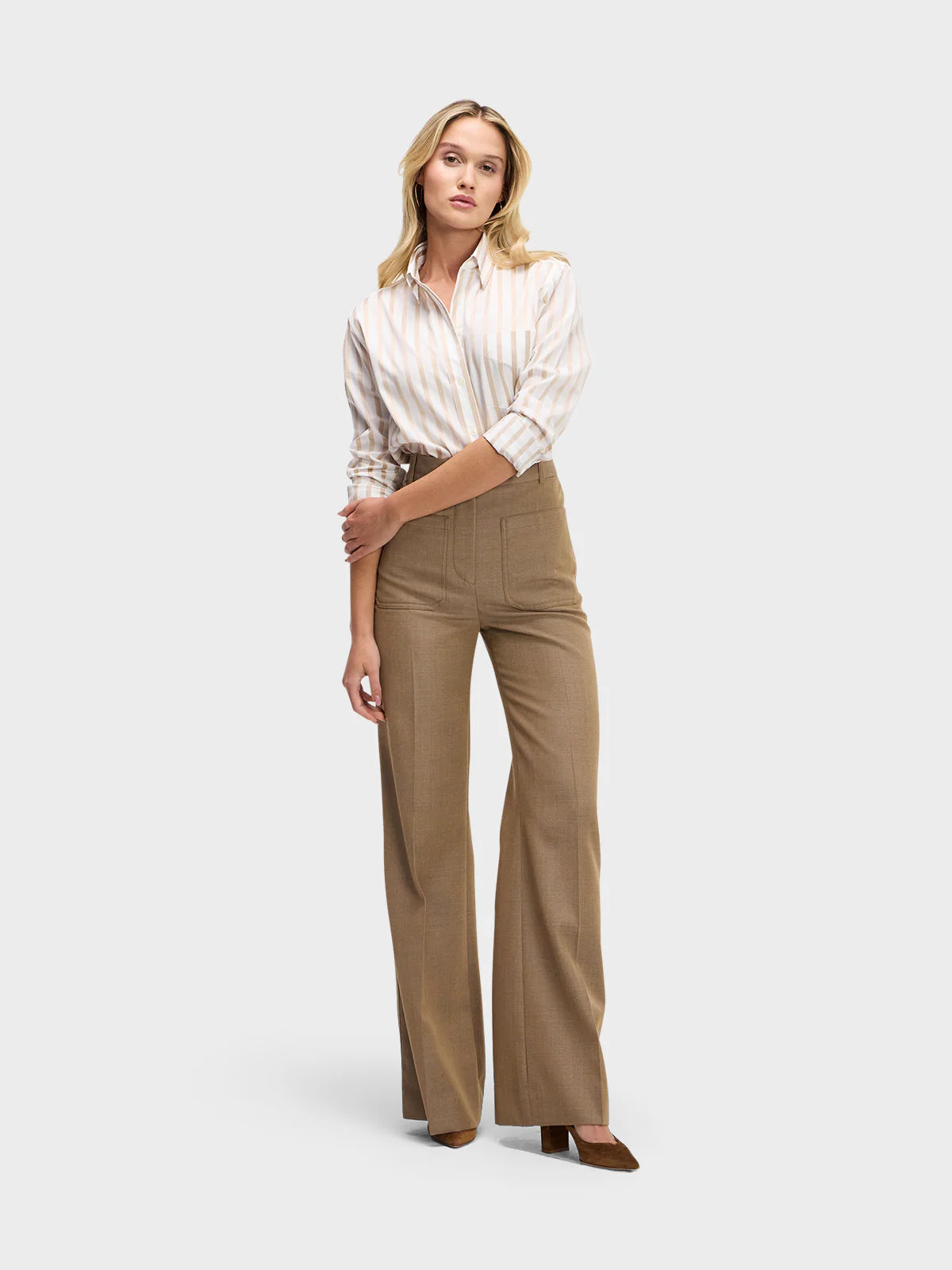 A woman stands against a plain background wearing the Weekend Shirt by Sarah Alexandra, LLC, a neutral striped top tucked into high-waisted, wide-leg brown trousers with front pockets and brown heels.