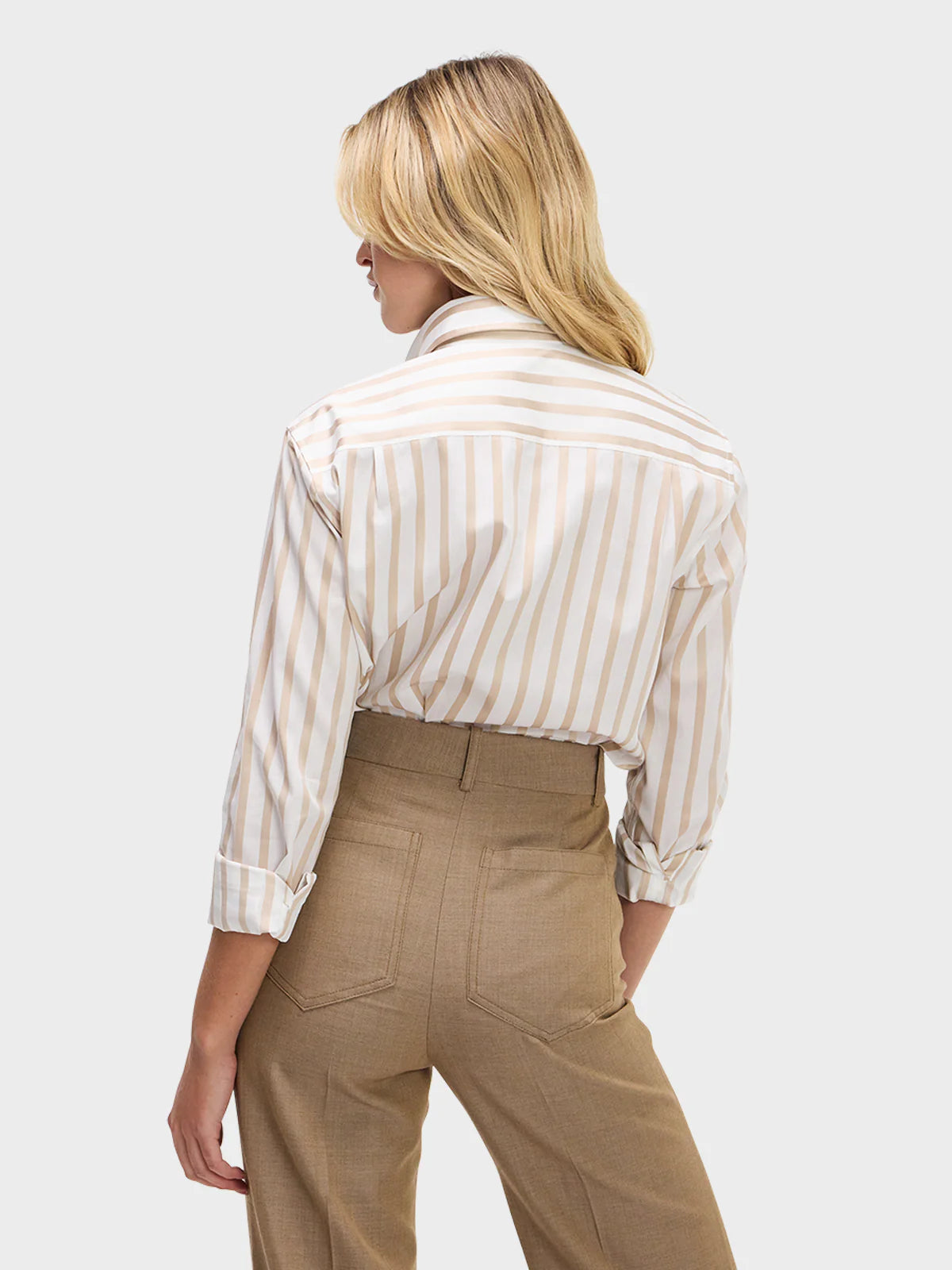 A woman with blonde hair, wearing the Sarah Alexandra Weekend Shirt and high-waisted light brown trousers, stands facing away from the camera against a plain light background.