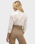 A woman with blonde hair, wearing the Sarah Alexandra Weekend Shirt and high-waisted light brown trousers, stands facing away from the camera against a plain light background.