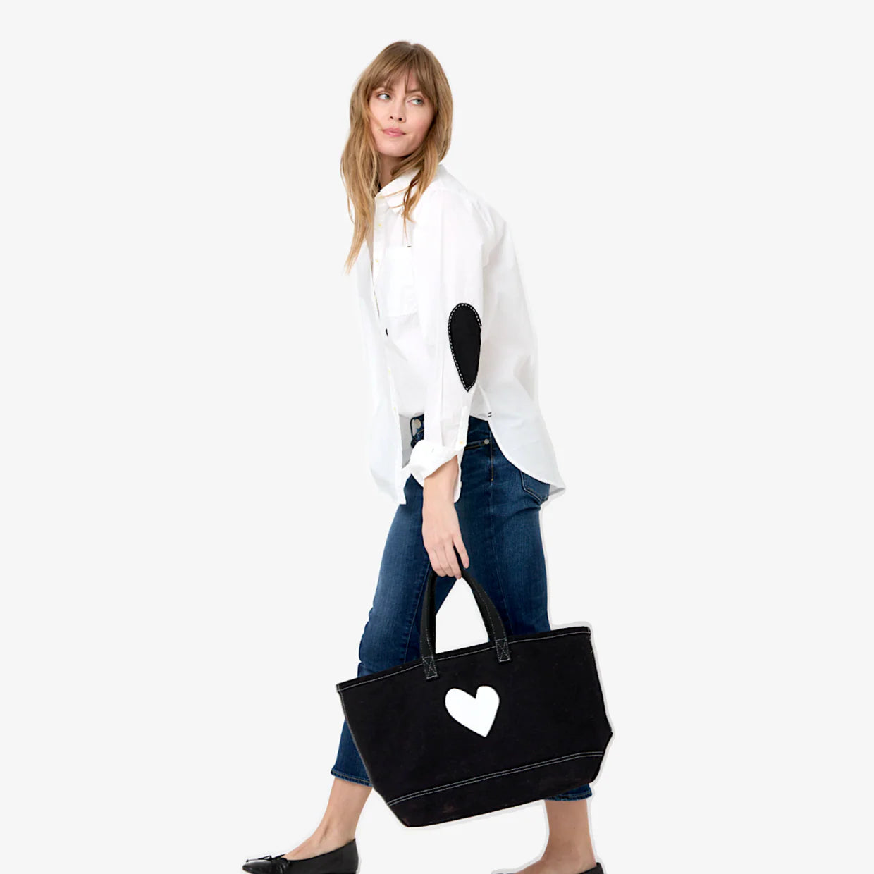 A woman wears the Kerri Rosenthal Mia Shirt Classic White with black elbow patches, blue jeans, and black flats, holding a large black tote bag with a white heart against a light background for a timeless wardrobe staple look.