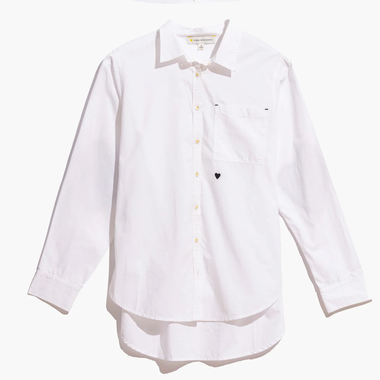 The Kerri Rosenthal Mia Shirt Classic White is a long-sleeve, crisp cotton poplin button-up with a chest pocket, small black heart embroidery nearby, and a subtle horizontal line detail—an essential wardrobe staple.