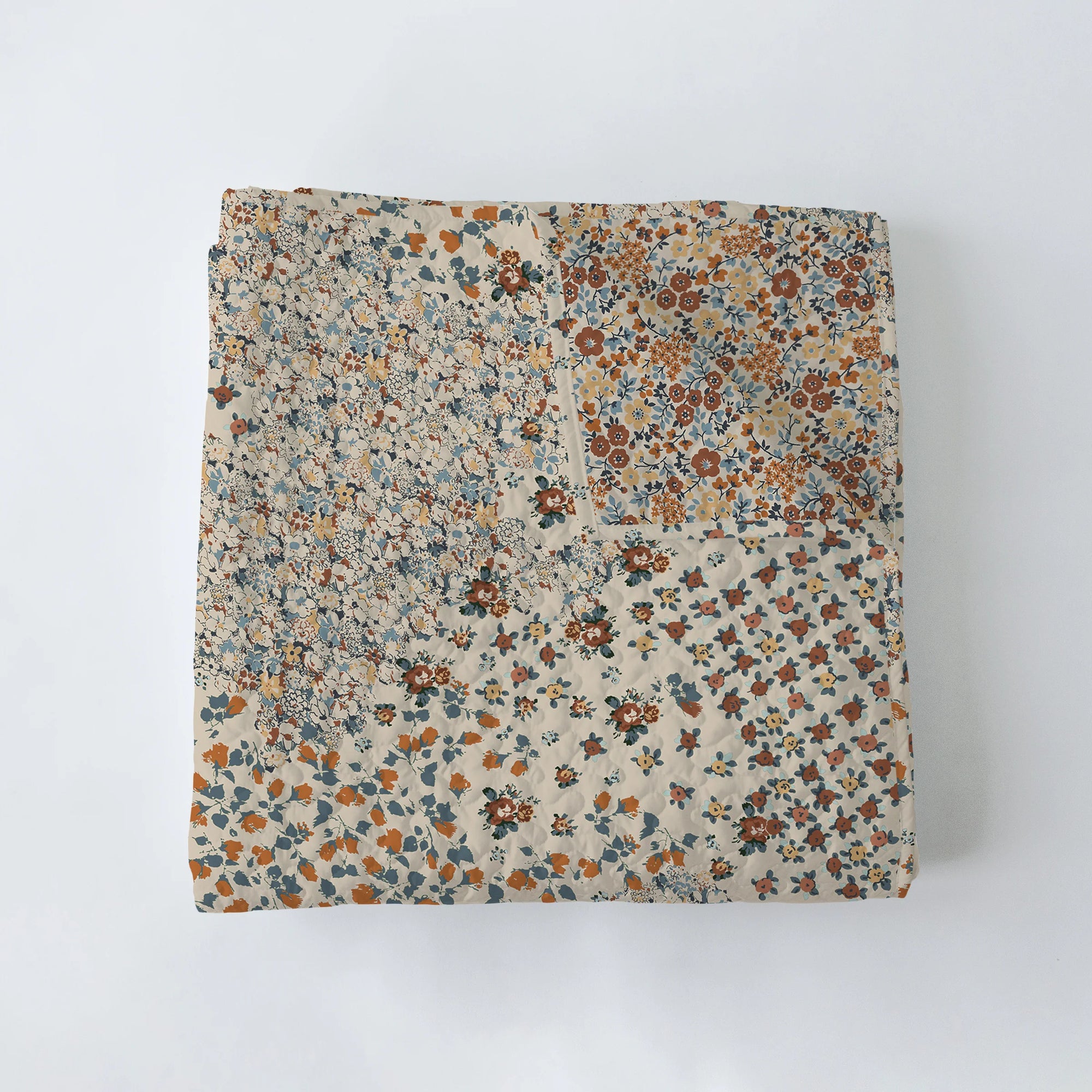 The Winchester Orange Quilt by Faire is a folded Texas-made patchwork quilt with small floral patterns in earthy brown, blue, orange, and beige tones, shown on a plain white background.