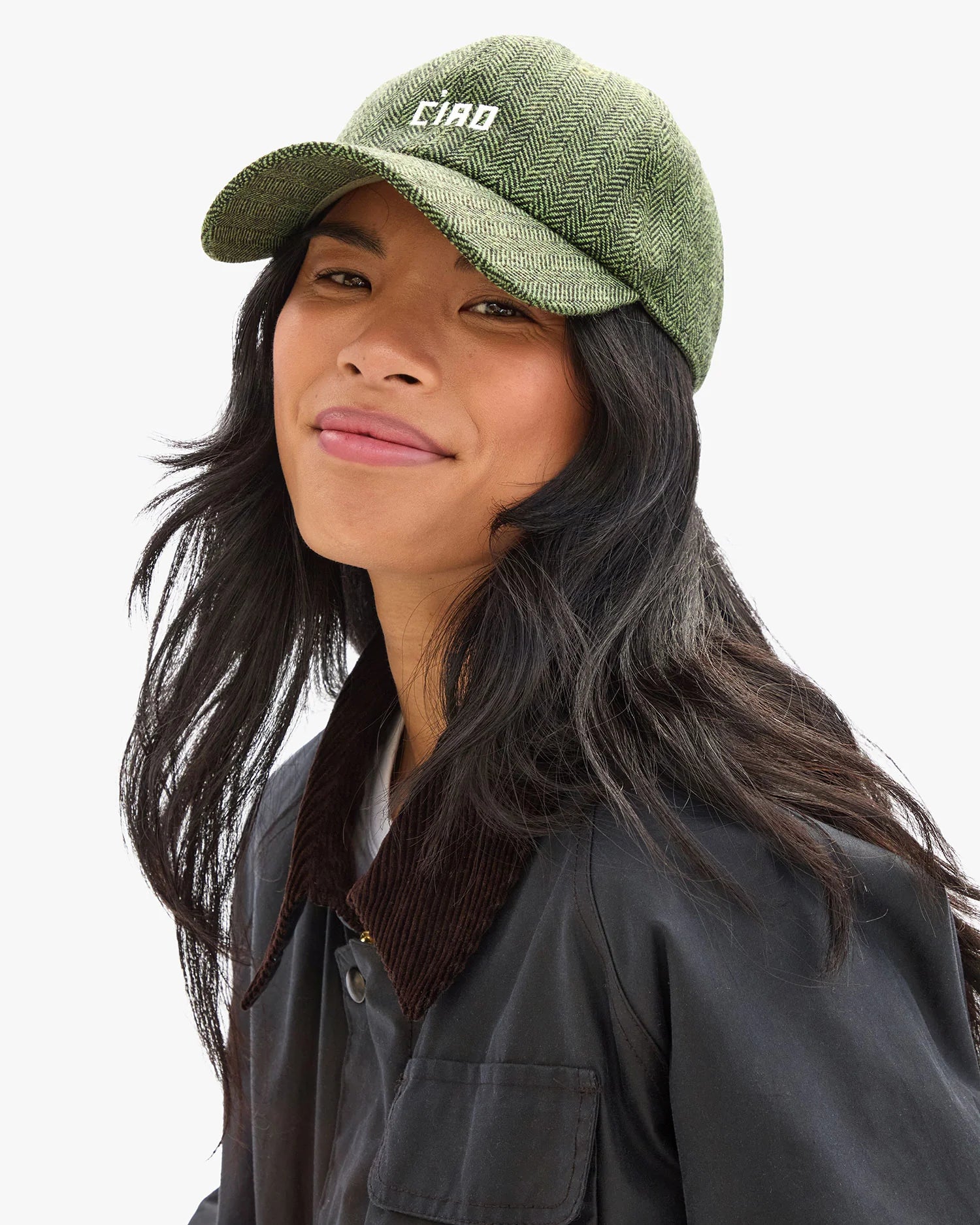 A person with long black hair wears the Clare Vivier Wool CIAO Baseball Hat—a green herringbone cap with "CIAO" embroidered on the front—and a dark jacket, smiling slightly against a white background.