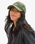 A person with long black hair wears the Clare Vivier Wool CIAO Baseball Hat—a green herringbone cap with "CIAO" embroidered on the front—and a dark jacket, smiling slightly against a white background.
