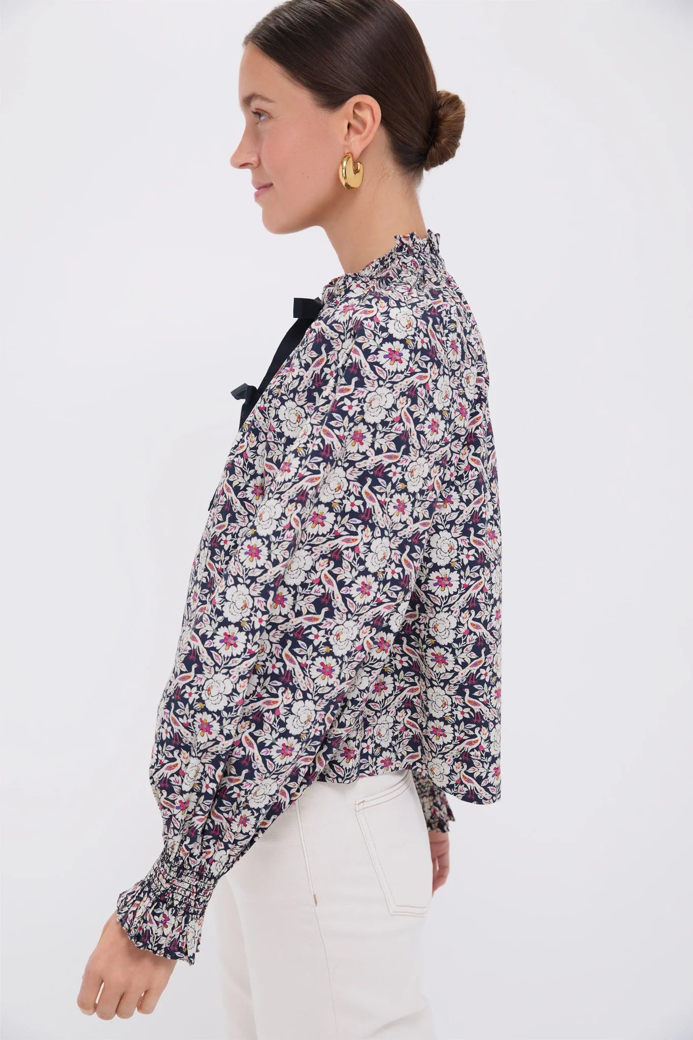 Against a plain white background, a person with dark hair pulled back and hoop earrings stands in profile wearing the Aiden Top by Rhode—a floral-patterned blouse with a ruffle collar and self-tie bows—paired with white pants.