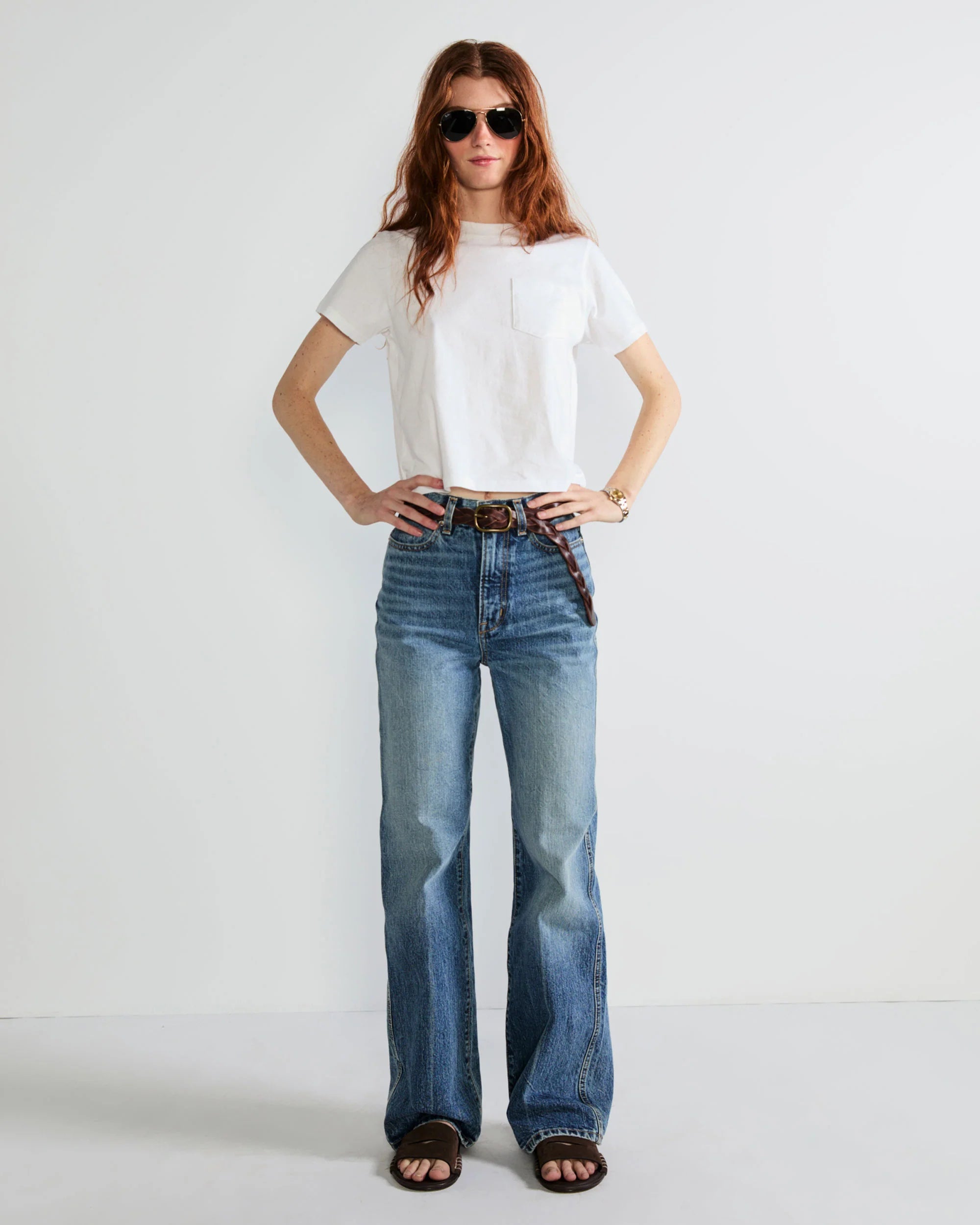 A woman with long red hair and sunglasses wears the Kule Wynn Baby Tee (white, 100% cotton), blue high-waisted jeans, a brown belt, and sandals, standing against a plain white background with hands on her hips.