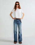 A woman with long red hair and sunglasses wears the Kule Wynn Baby Tee (white, 100% cotton), blue high-waisted jeans, a brown belt, and sandals, standing against a plain white background with hands on her hips.