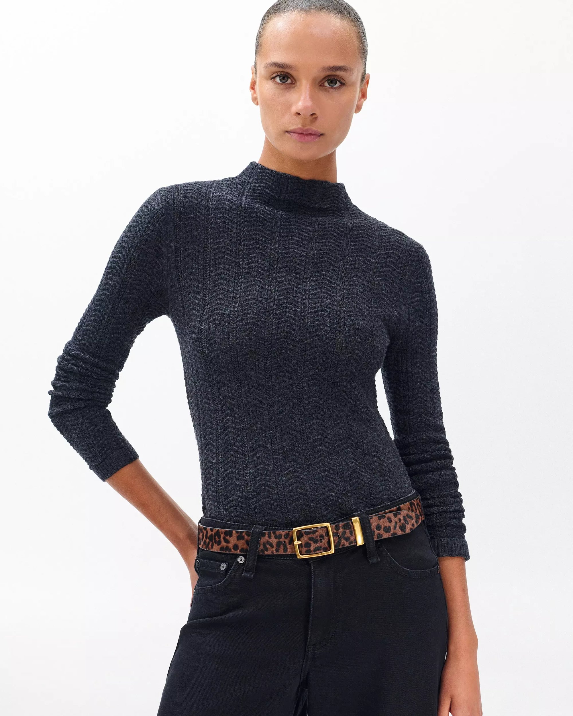 A woman with slicked-back hair wears a textured black long-sleeve top, black jeans, and the Rag & Bone X Boyfriend Belt, standing against a plain white background.