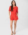 A woman with long brown hair and a neutral expression wears the Xirena Evert Romper—a bright red cotton-blend terry romper with button neckline—posing against a white background, her left hand in her pocket, paired with beige flat sandals.