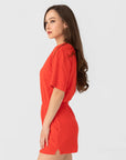 A woman with long brown hair, wearing the Xirena Evert Romper in bright red towel terry, stands sideways against a plain light gray background.