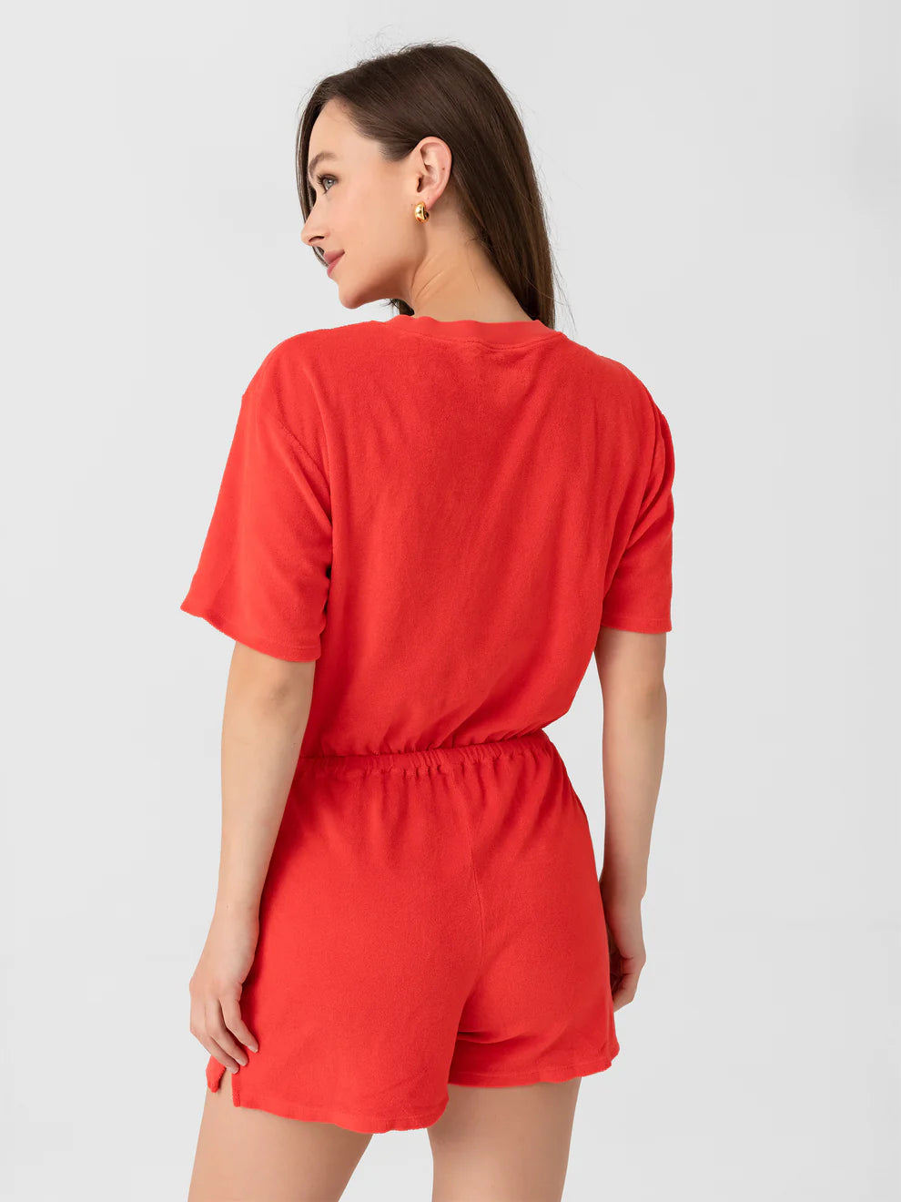 A woman with long brown hair models the Xirena Evert Romper in bright red cotton-blend terry, shown from the back as she glances over her shoulder and smiles, accessorized with gold hoop earrings against a plain light background.