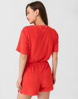 A woman with long brown hair models the Xirena Evert Romper in bright red cotton-blend terry, shown from the back as she glances over her shoulder and smiles, accessorized with gold hoop earrings against a plain light background.