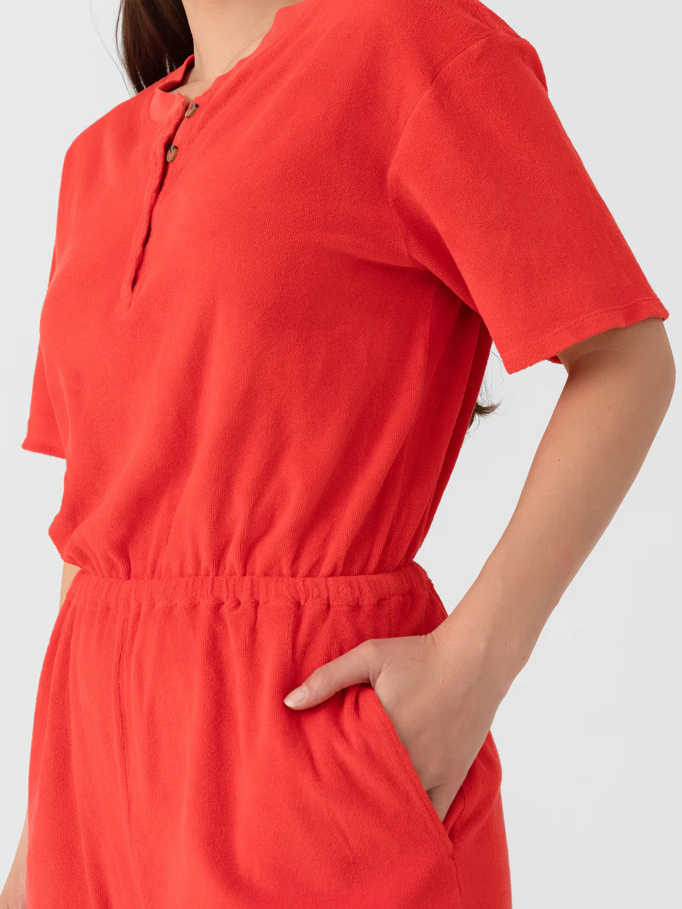 A person wears the Xirena Evert Romper, a bright red cotton-blend terry piece with short sleeves and matching high-waisted shorts featuring an elastic waistband, shown from shoulders to thighs with one hand in a pocket.