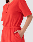 A person wears the Xirena Evert Romper, a bright red cotton-blend terry piece with short sleeves and matching high-waisted shorts featuring an elastic waistband, shown from shoulders to thighs with one hand in a pocket.
