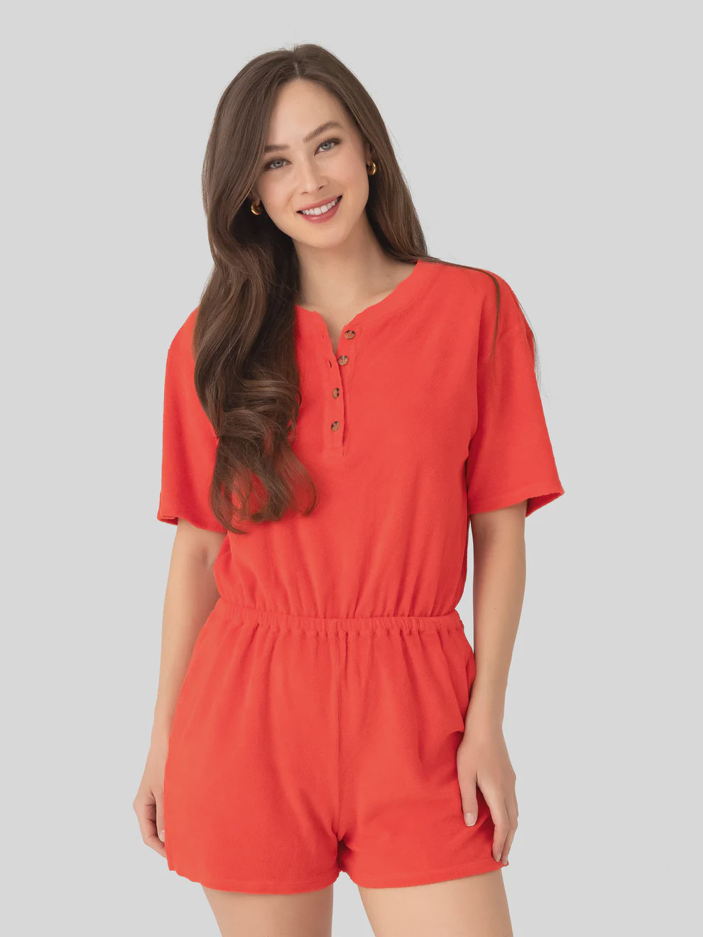 A woman with long brown hair is smiling and wearing the Xirena Evert Romper, a bright orange cotton-blend towel terry piece with an elastic waistband, standing against a light gray background.