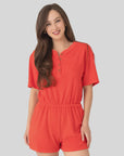 A woman with long brown hair is smiling and wearing the Xirena Evert Romper, a bright orange cotton-blend towel terry piece with an elastic waistband, standing against a light gray background.