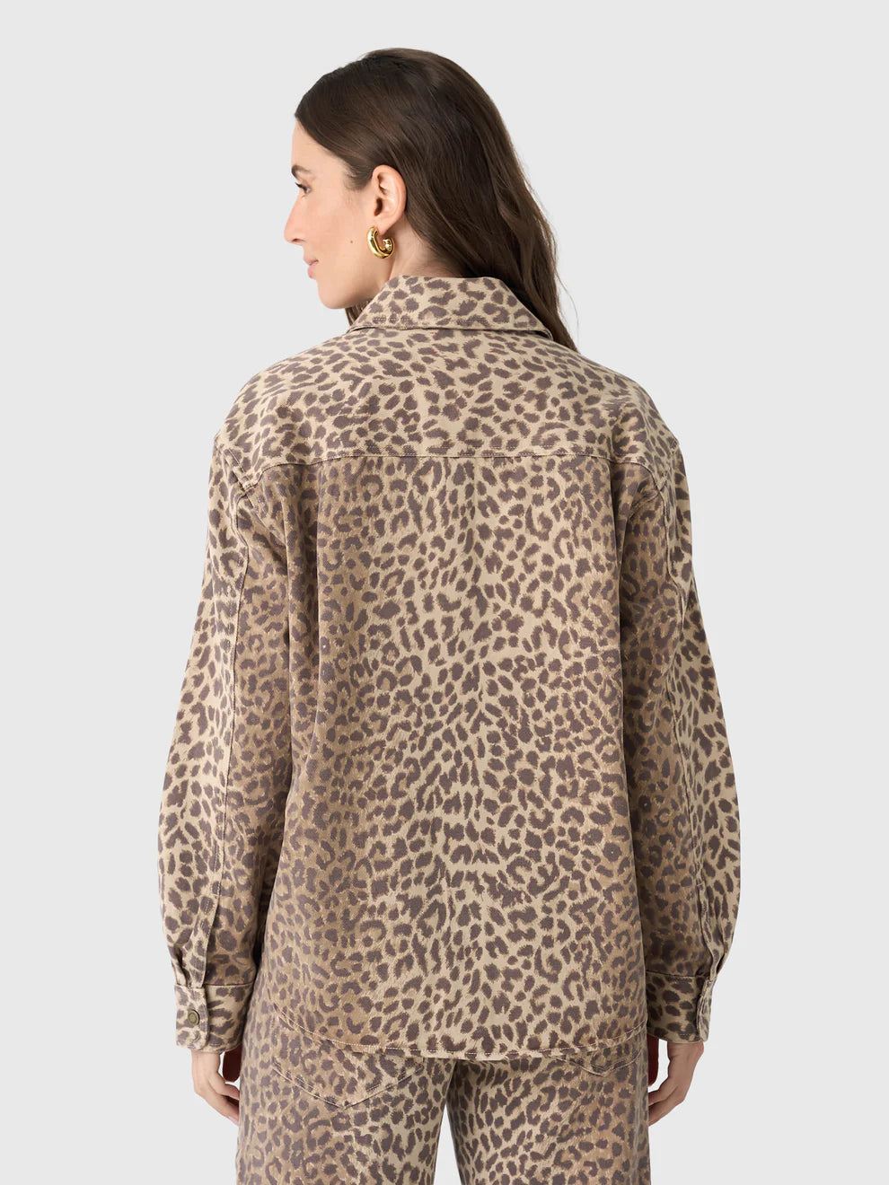 A woman with long brown hair and gold hoop earrings models the Xirena Gia Shirt, a relaxed fit beige and brown leopard print button-up with a spread collar, paired with matching pants, set against a plain light background.