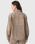 A woman with long brown hair and gold hoop earrings models the Xirena Gia Shirt, a relaxed fit beige and brown leopard print button-up with a spread collar, paired with matching pants, set against a plain light background.