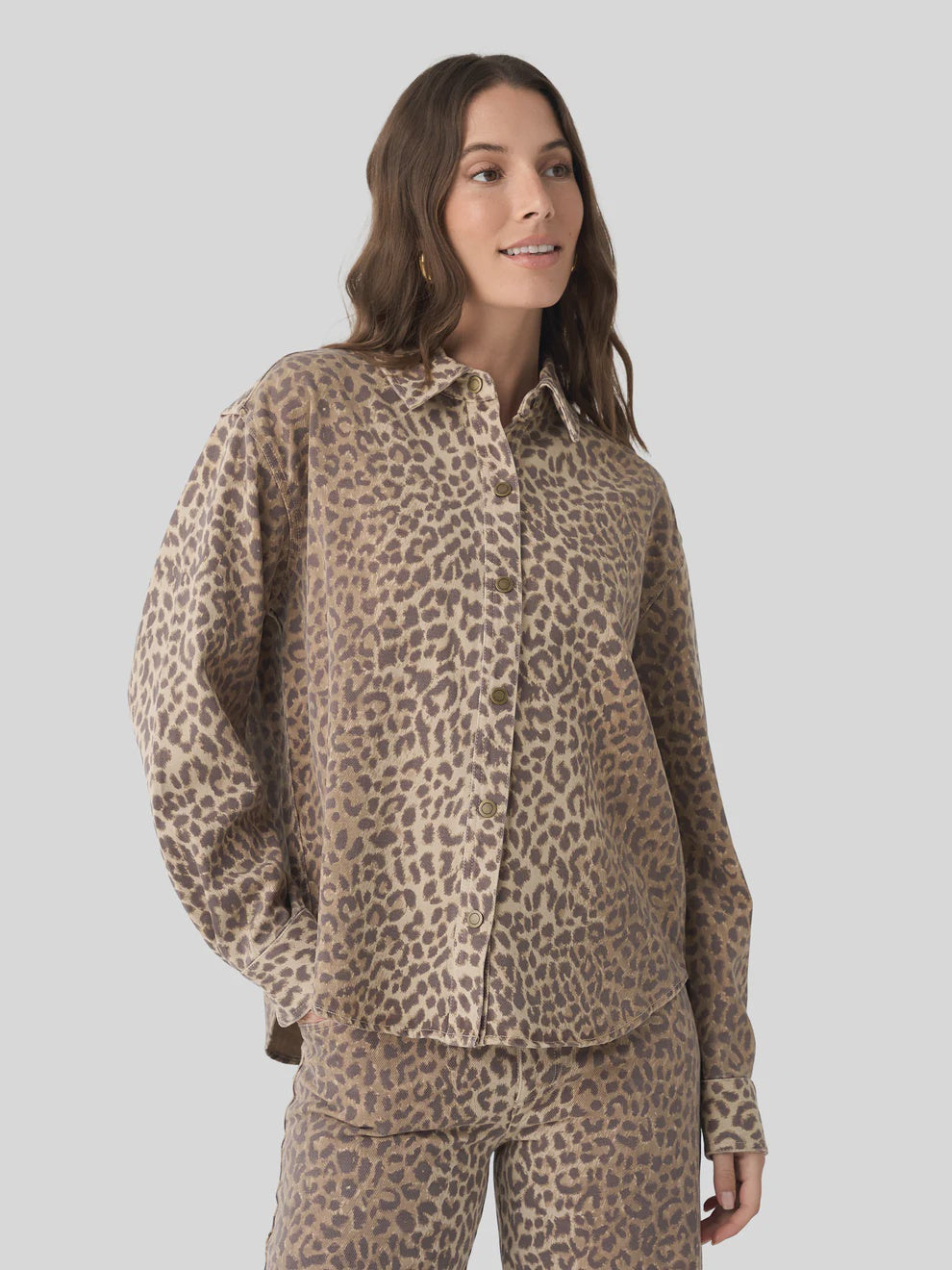 A woman with long brown hair wears the Xirena Gia Shirt—a relaxed fit, beige and brown leopard print button-up with a spread collar—paired with matching pants, standing against a light gray background and looking to the side, smiling.
