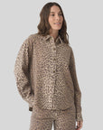 A woman with long brown hair wears the Xirena Gia Shirt—a relaxed fit, beige and brown leopard print button-up with a spread collar—paired with matching pants, standing against a light gray background and looking to the side, smiling.