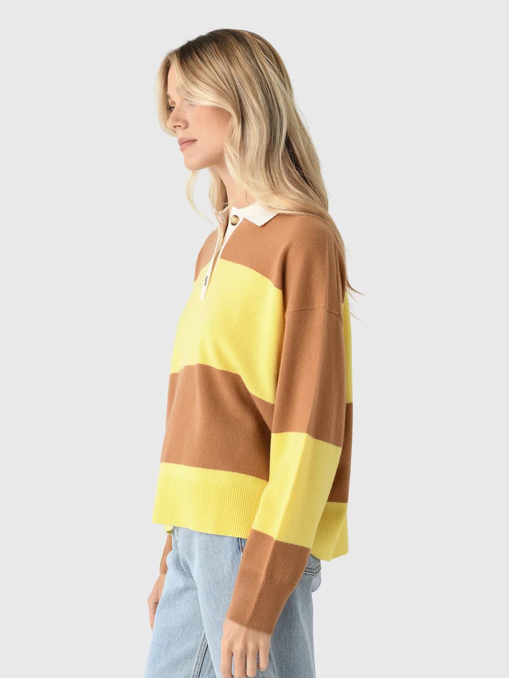 A woman with long blonde hair wears the Xirena Braxton Sweater— a loose, polo-style knit with wide yellow and brown stripes—paired with light blue jeans, standing against a plain light gray background.