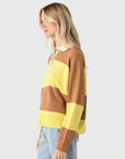 A woman with long blonde hair wears the Xirena Braxton Sweater— a loose, polo-style knit with wide yellow and brown stripes—paired with light blue jeans, standing against a plain light gray background.