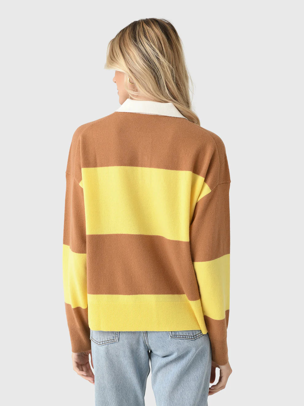 A woman with long blonde hair, seen from behind, wears the Xirena Braxton Sweater—featuring brown and yellow stripes with a white collar—and light blue jeans against a plain light gray background.