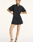 A woman with loose waves poses with one hand on her hip, wearing the Xirena Franklin Romper—a short-sleeved black button-up with utility patch pockets—and black strappy sandals against a plain background.