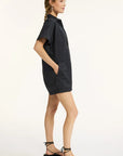 A woman stands in profile wearing the Xirena Franklin Romper, a collared button-down with front pockets for subtle utility. She pairs it with black strappy flat sandals, her wavy hair in a braid, set against a plain light background.