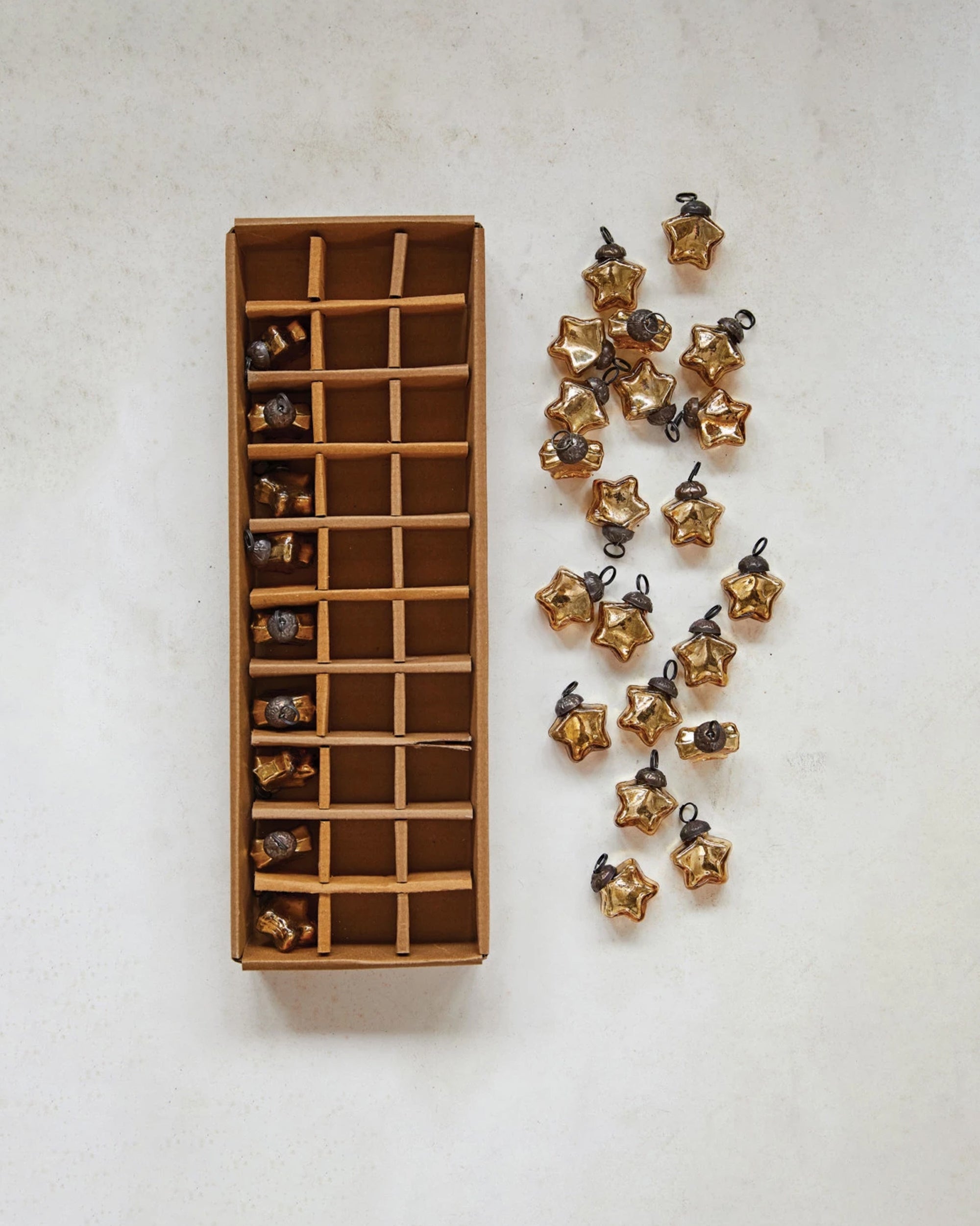 A cardboard box from Creative Co-op holds twenty-four Mini Glass Star Ornaments, each gold-finished. Eighteen similar ornaments are scattered nearby on a white surface.