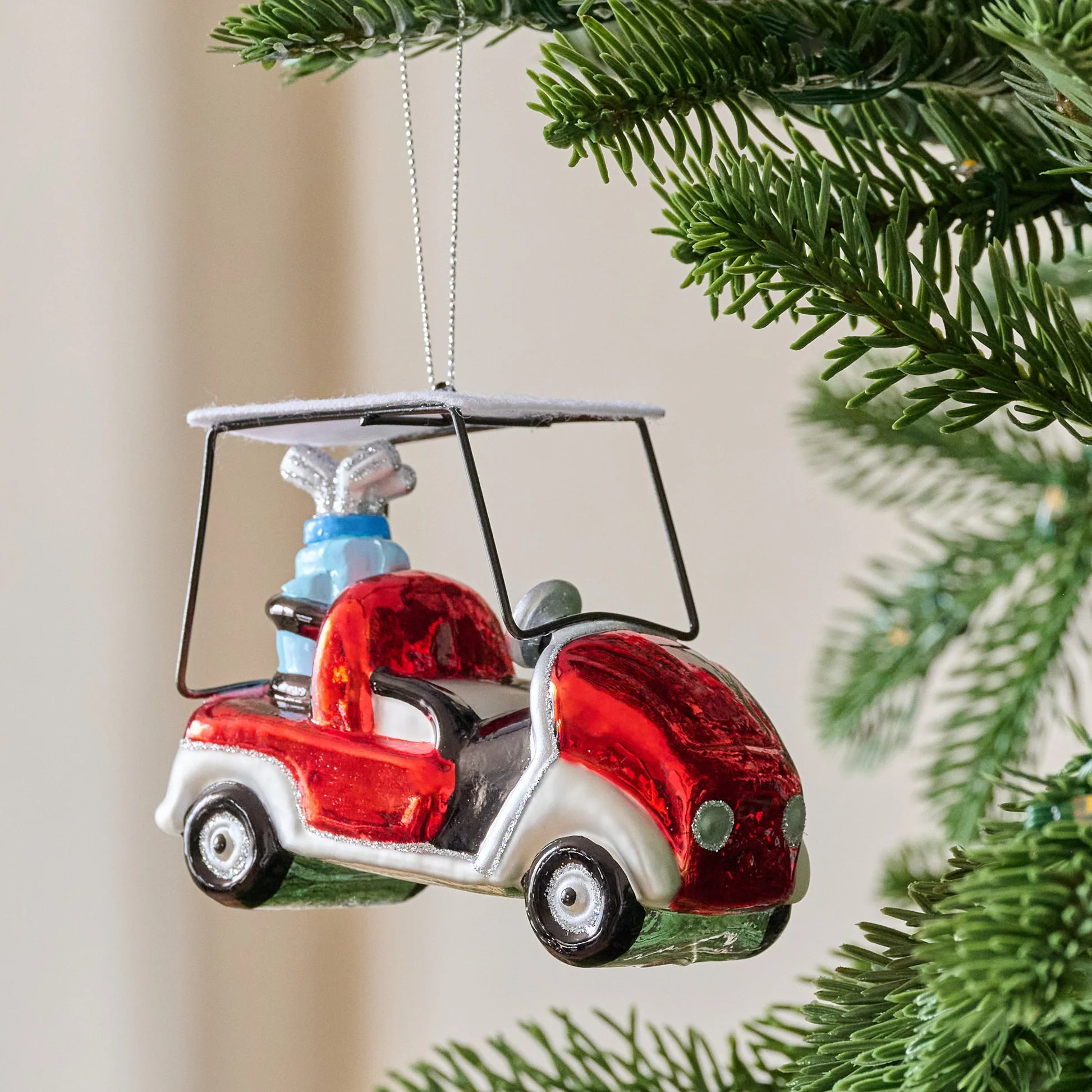 The Creative Co-op Glass Golf Cart Ornament, shaped like a red and white cart with a silver roof and a blue present in the back seat, is perfect for golf fans and makes a festive addition to your Christmas tree.