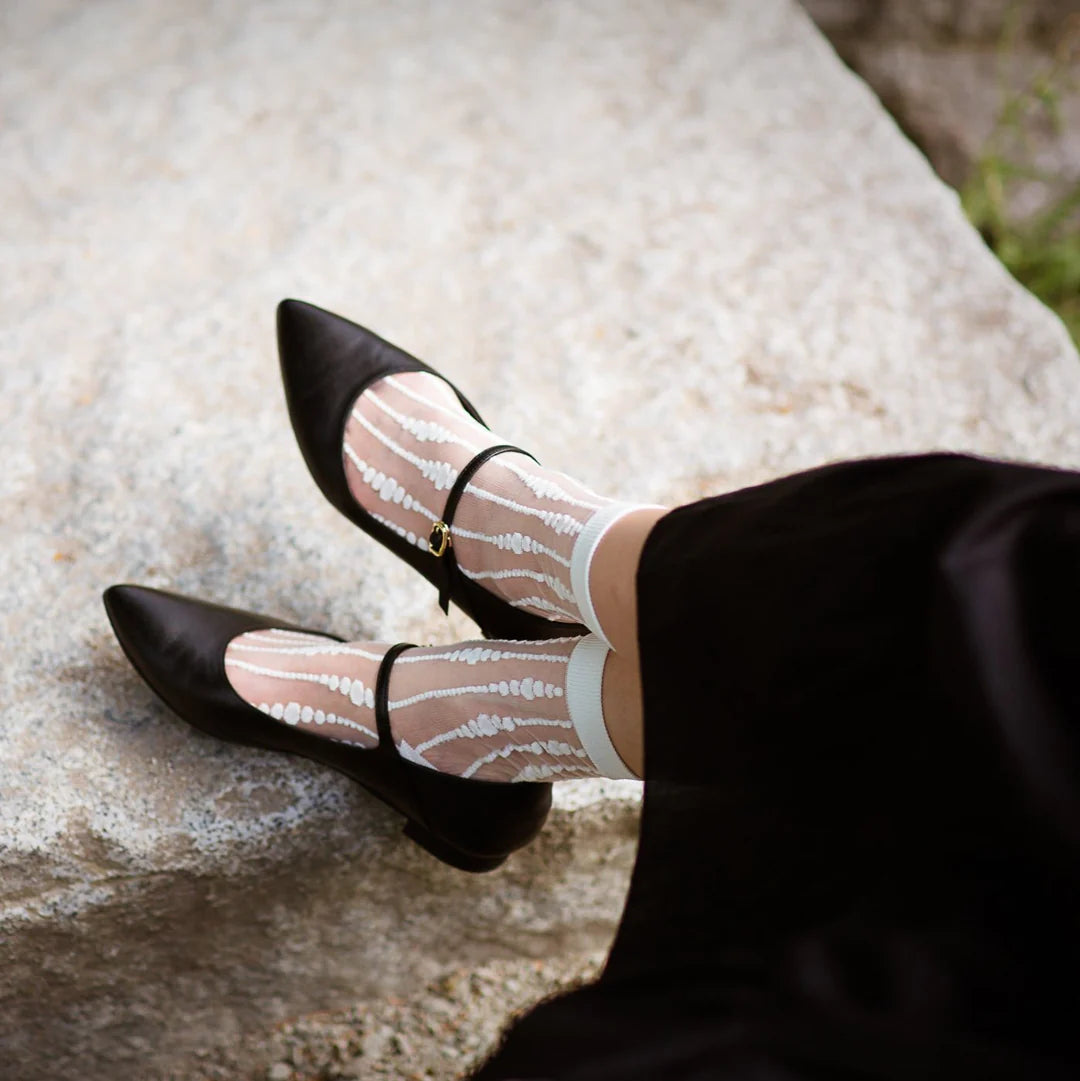 A person in a black skirt sits on a light stone surface, wearing Unified Sheer Bubble Socks and black Mary Jane shoes.